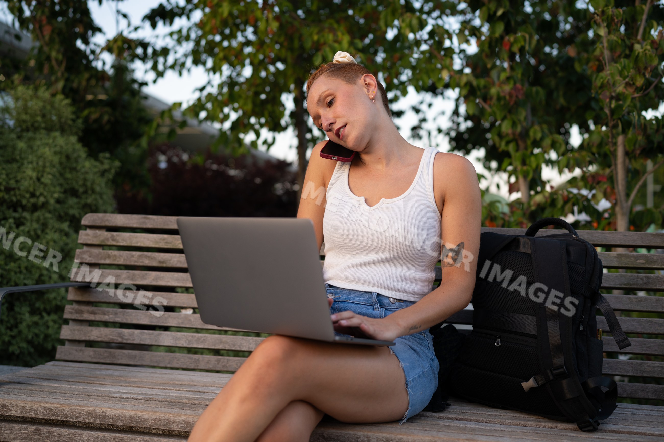 White Woman multitasks with laptop and phone on outdoor bench
