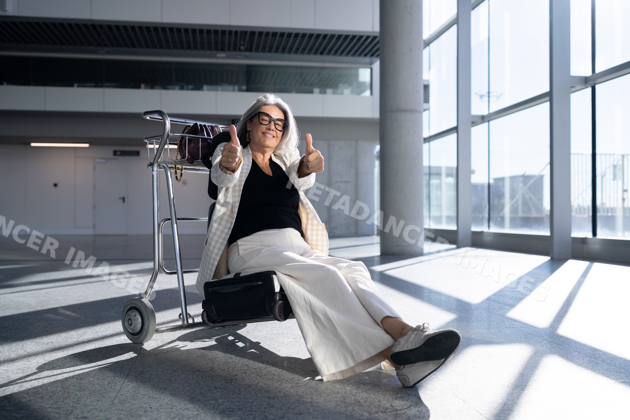 White woman traveler showing thumbs up sitting on luggage trolley