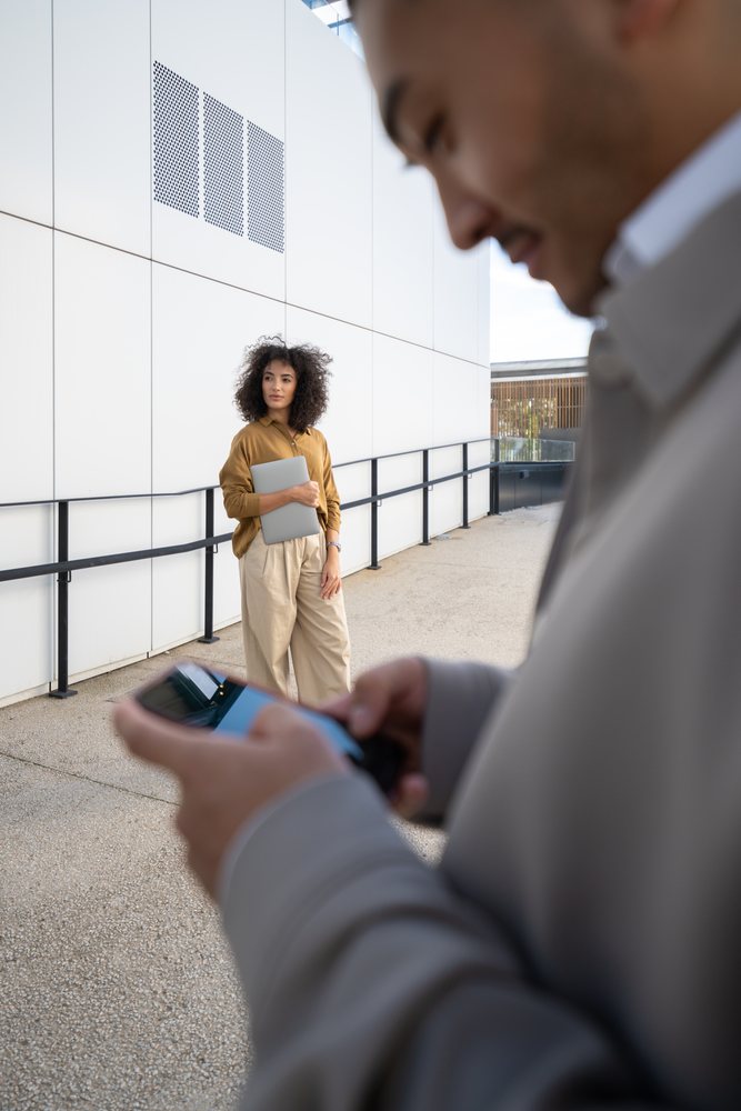 Woman holding laptop outside near building with man on smartphone