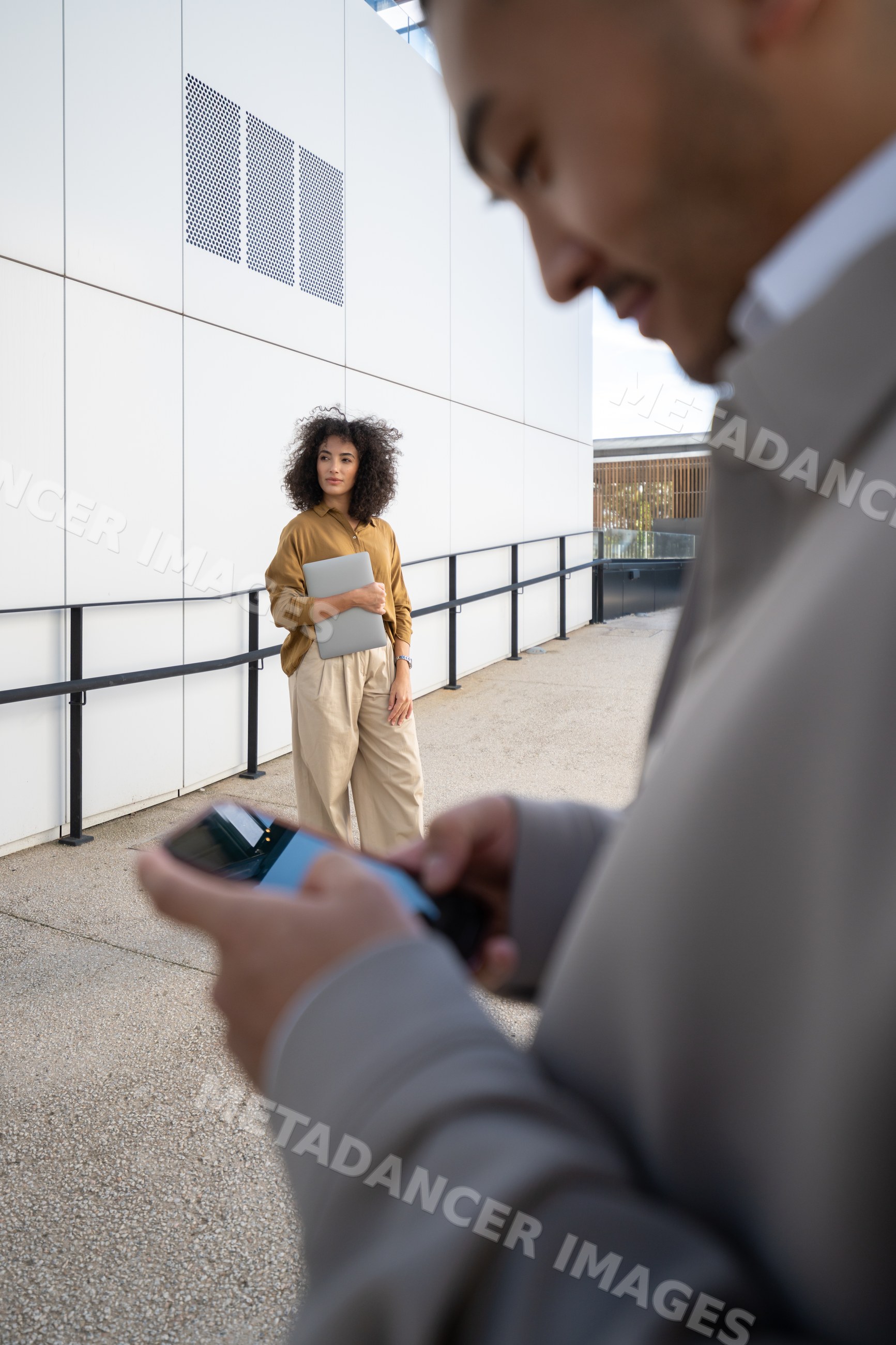 Woman holding laptop outside near building with man on smartphone