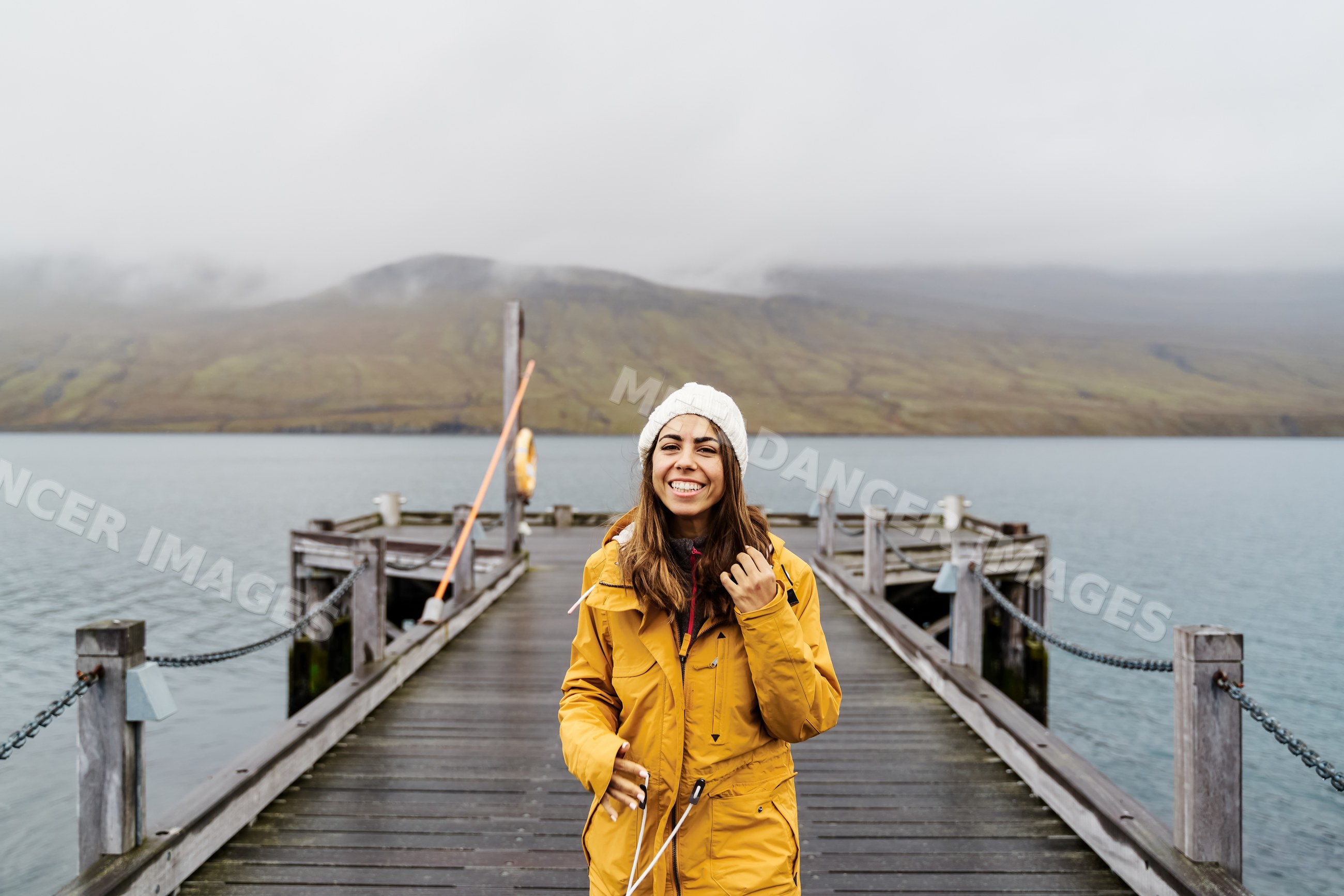 A white woman in a yellow jacket smiles on a wooden pier by water