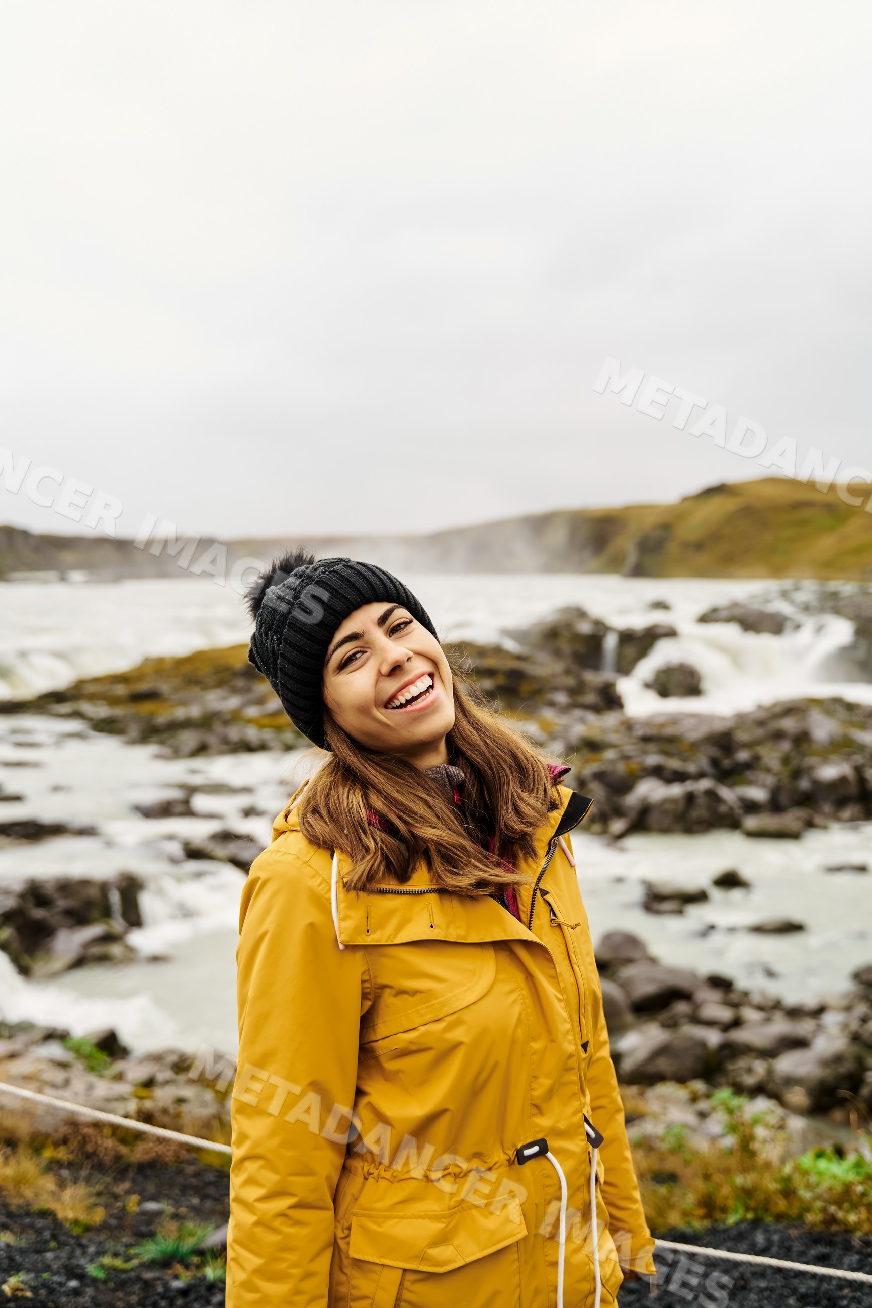 A white woman in a yellow jacket smiling outdoors near a waterfall