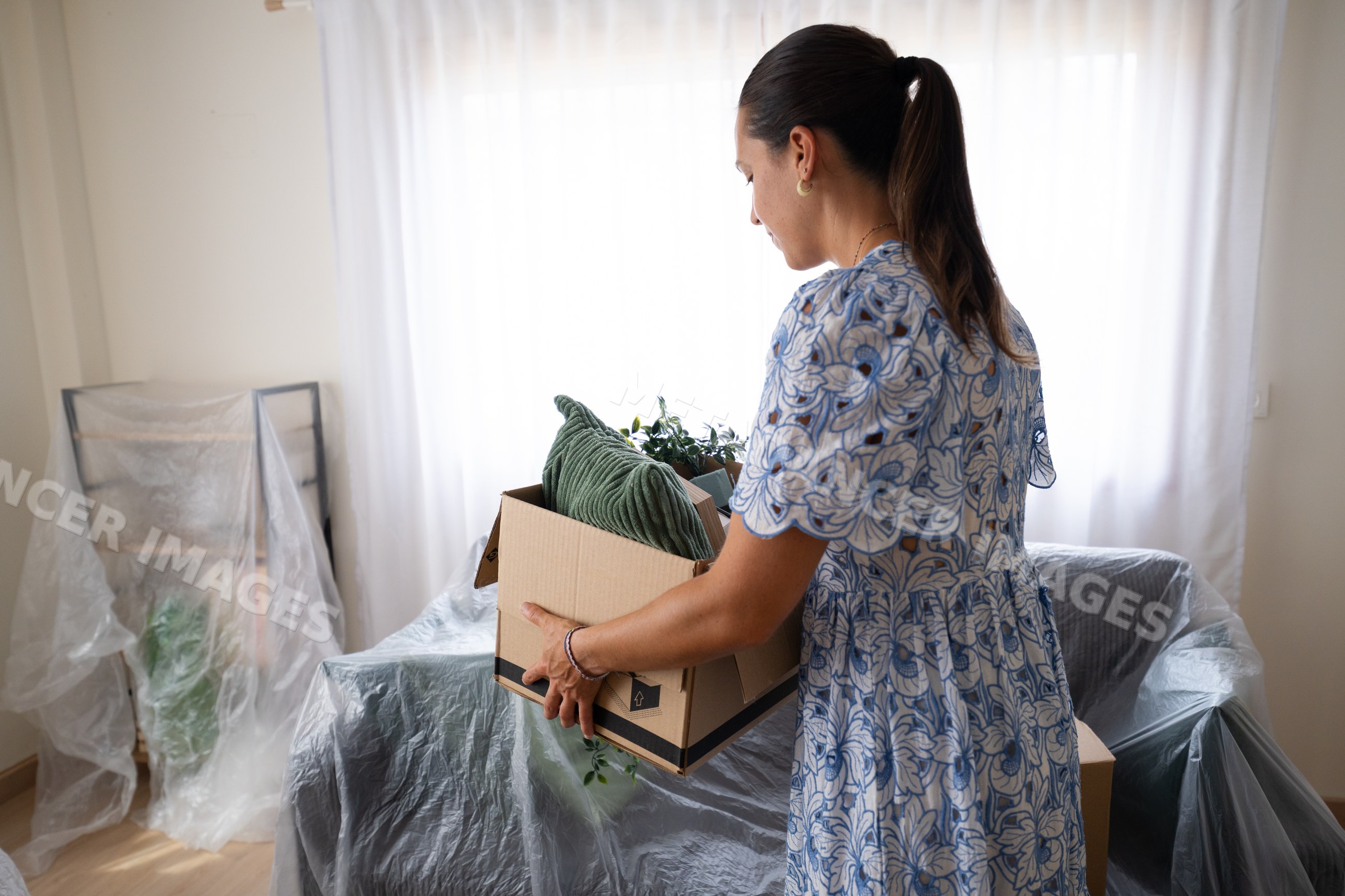 White woman carrying box with houseplants and cushion