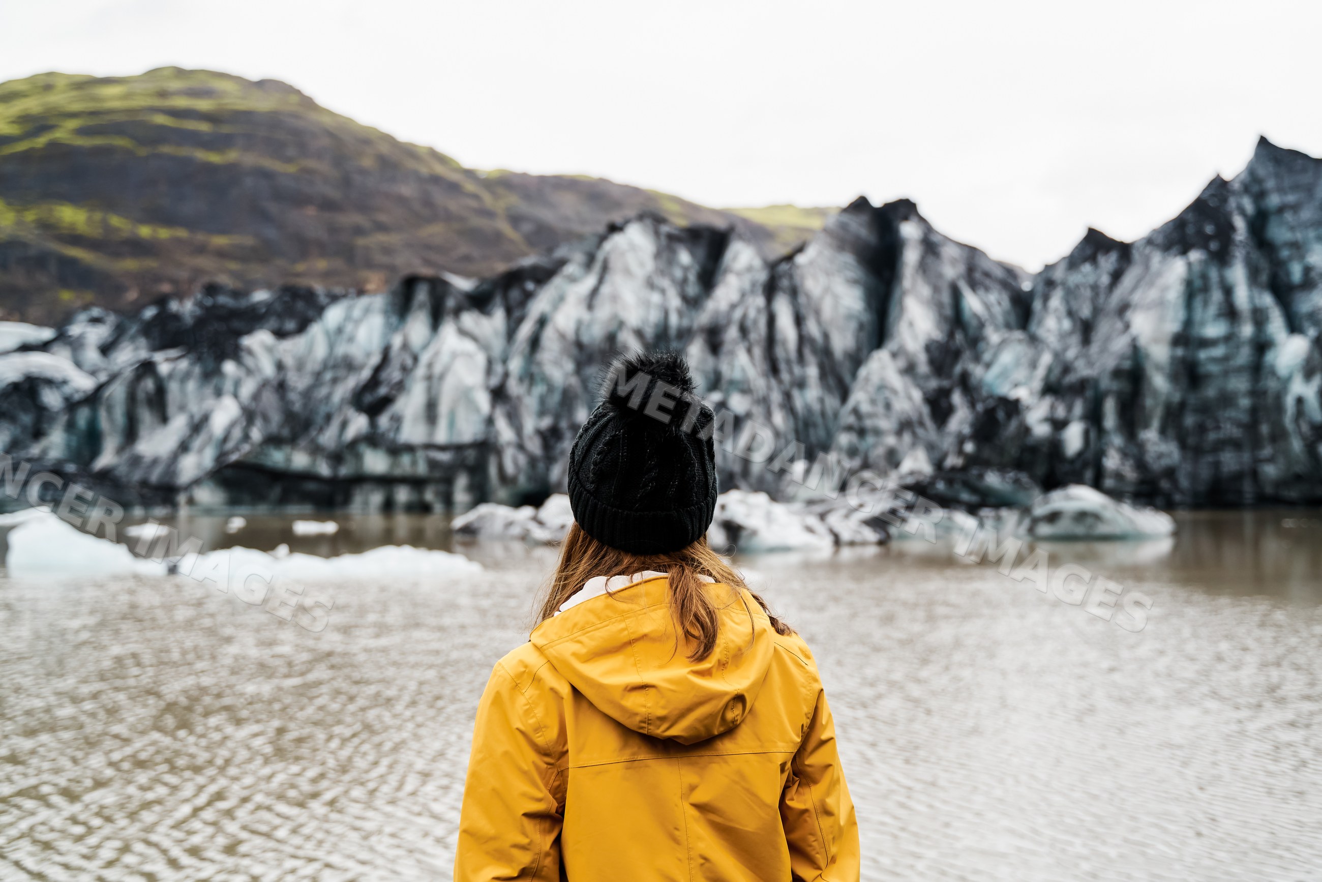 A white person wearing a yellow jacket looks out over a glacier lake