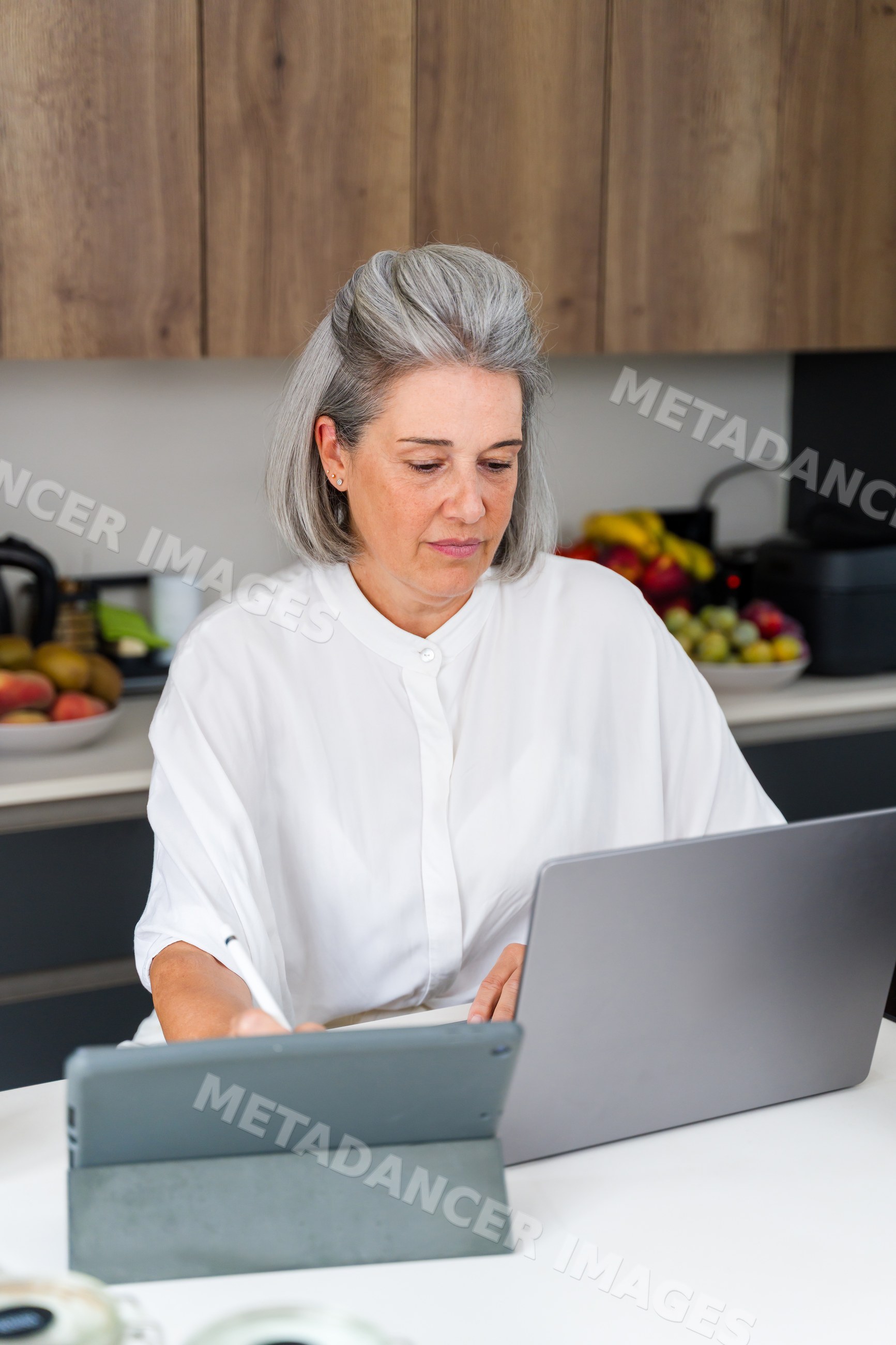 Middle aged white woman using laptop and tablet in modern home kitchen