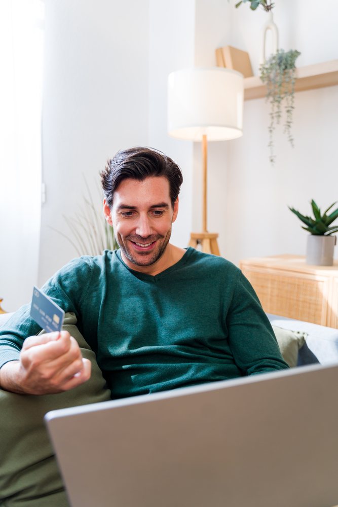 White man holding credit card using laptop in living room