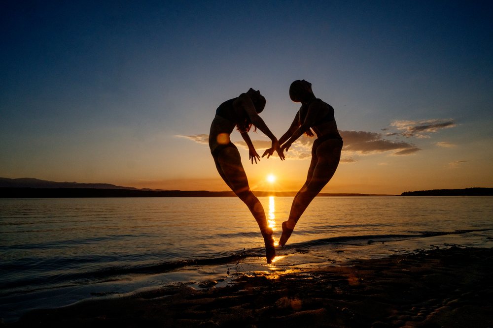 Two white people doing partner acrobatics jumps on a beach at sunset