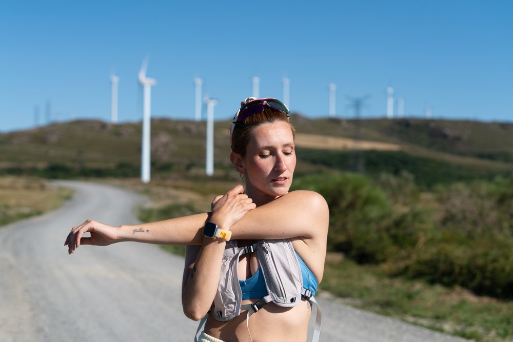 A white woman stretches her arm on a rural trail near wind turbines