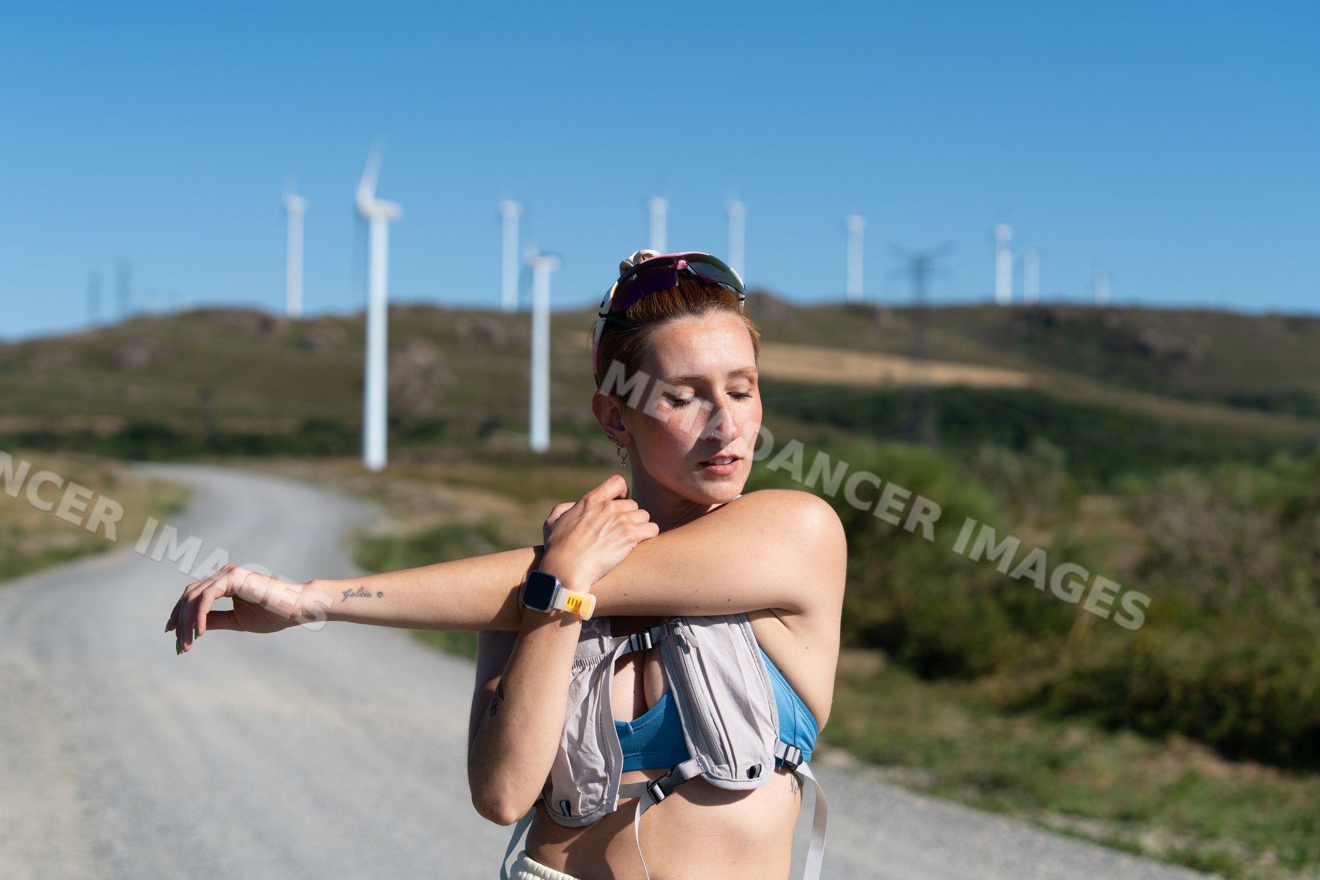A white woman stretches her arm on a rural trail near wind turbines
