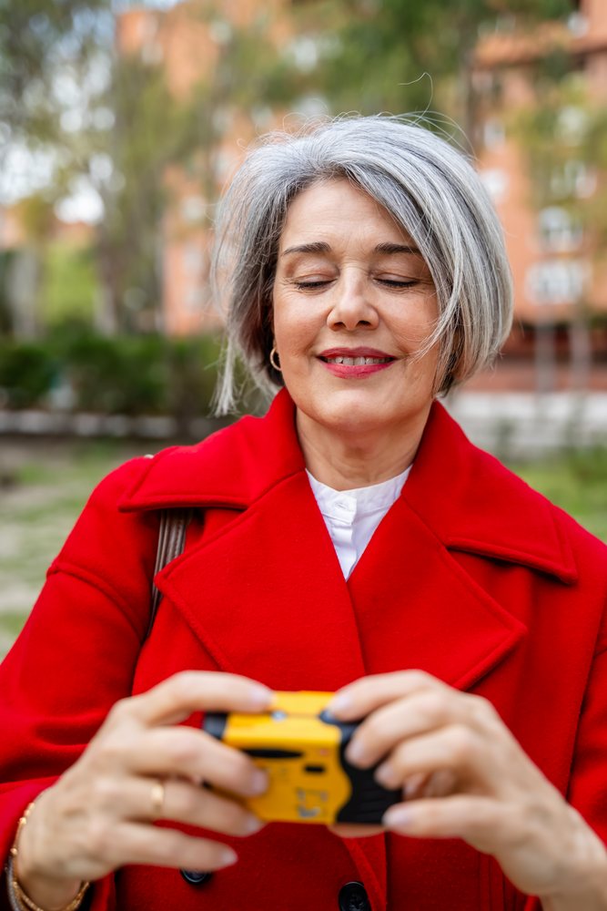 White woman with gray hair holding a small yellow camera outdoors