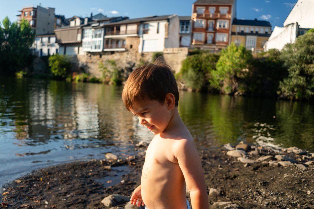 Shirtless white boy stands by the riverbank with buildings
