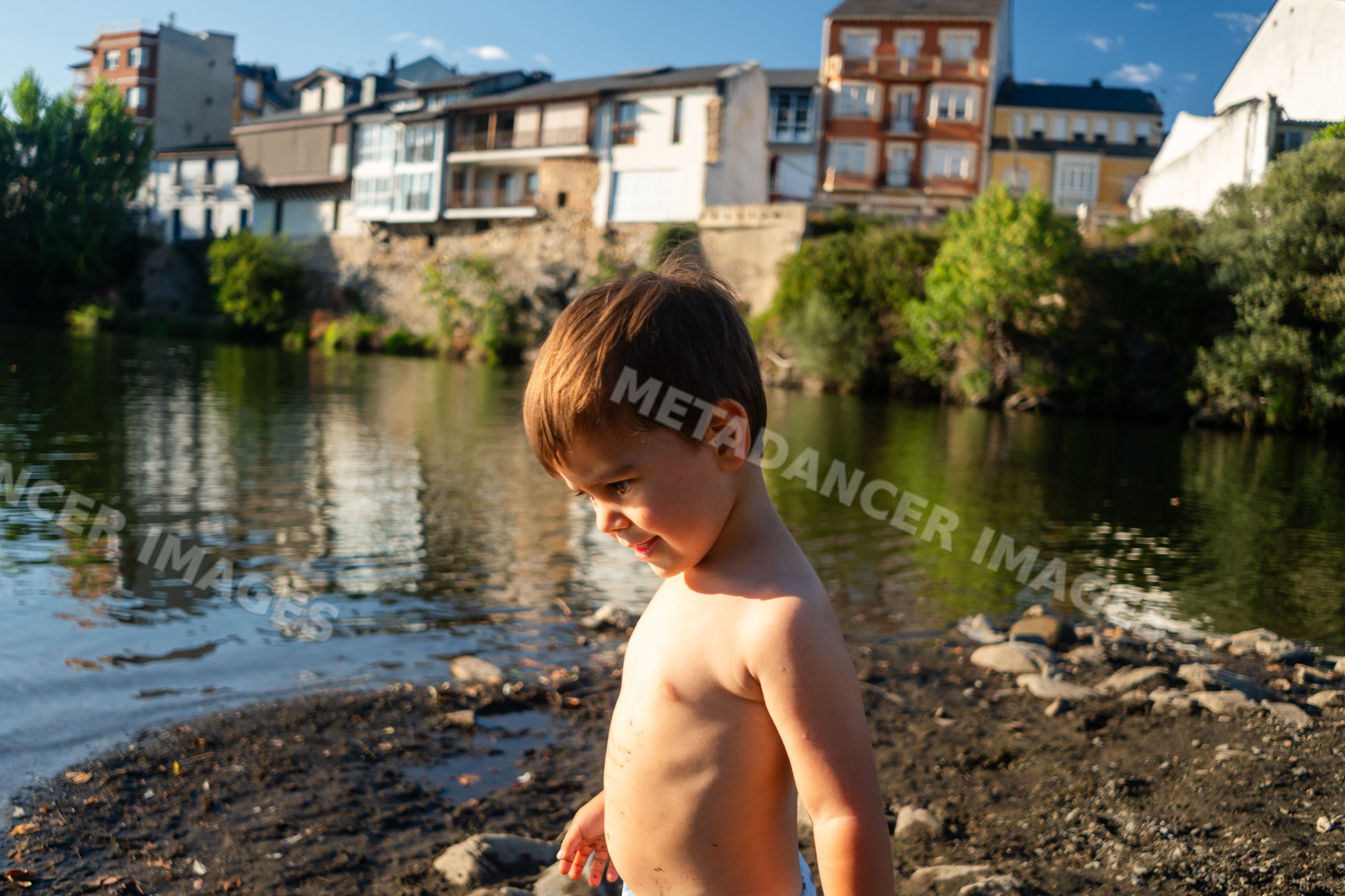 Shirtless white boy stands by the riverbank with buildings