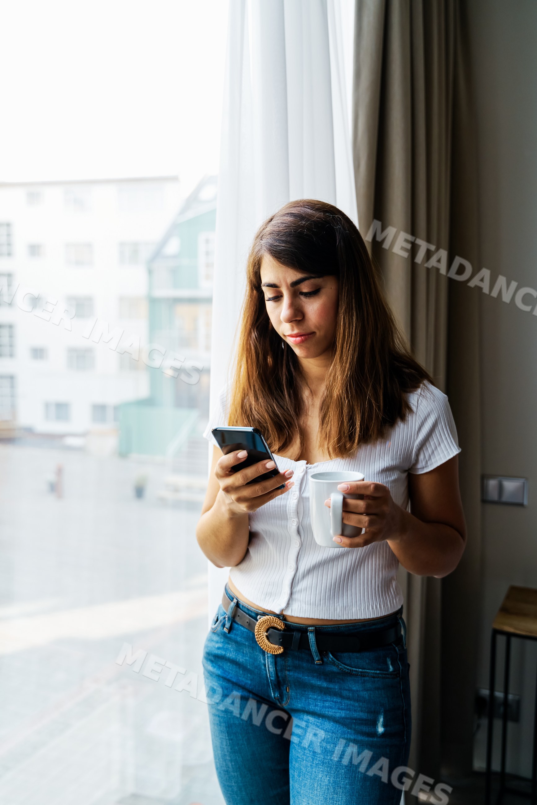 Woman using smartphone and holding cup indoors near large window