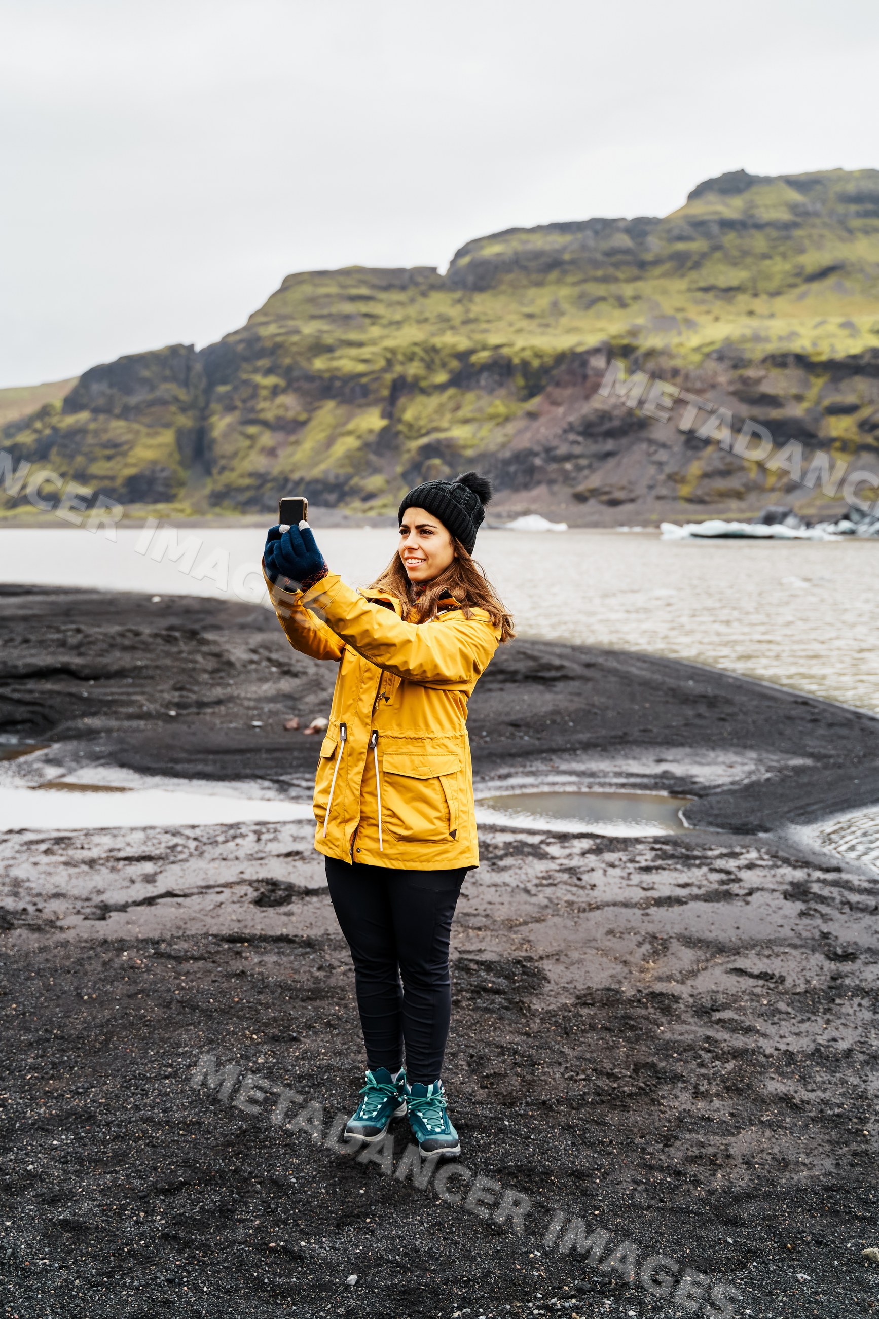 A white woman takes a selfie on volcanic black sand by a lake