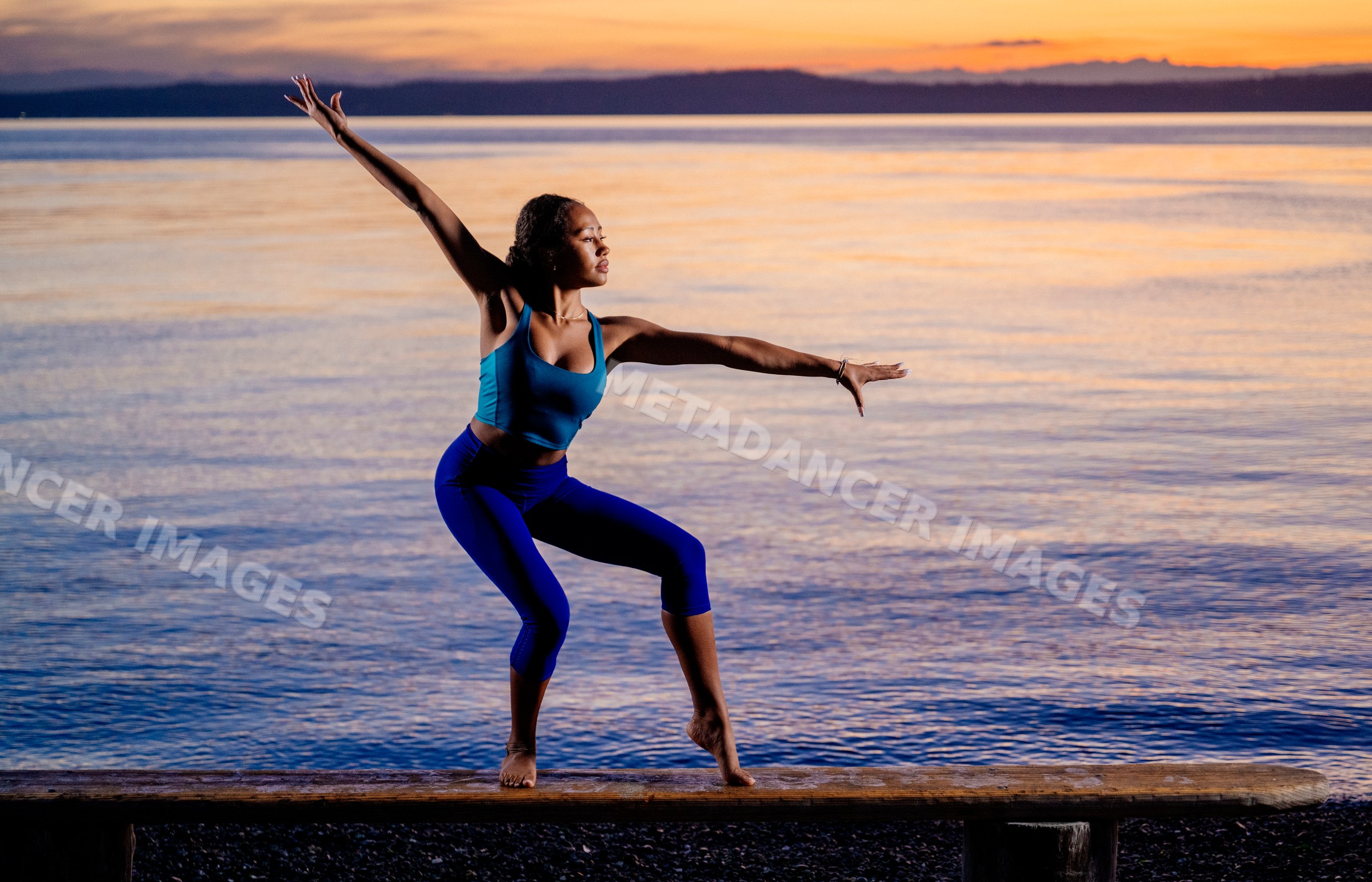 Hispanic woman balancing in yoga pose on wooden beam by lake at sunset