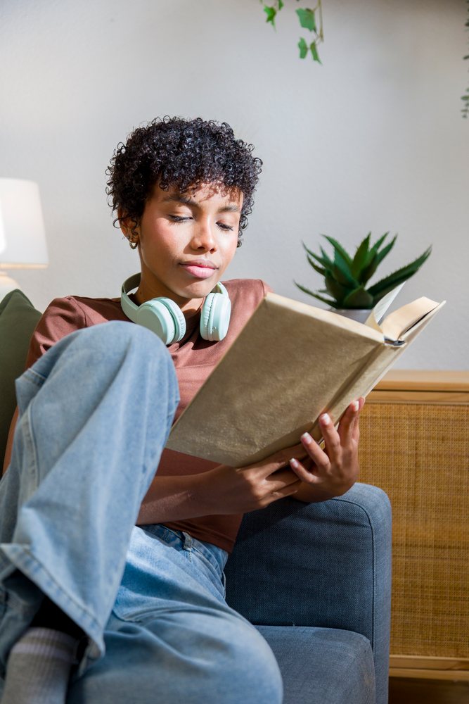 Young African American woman reading book with headphones on couch
