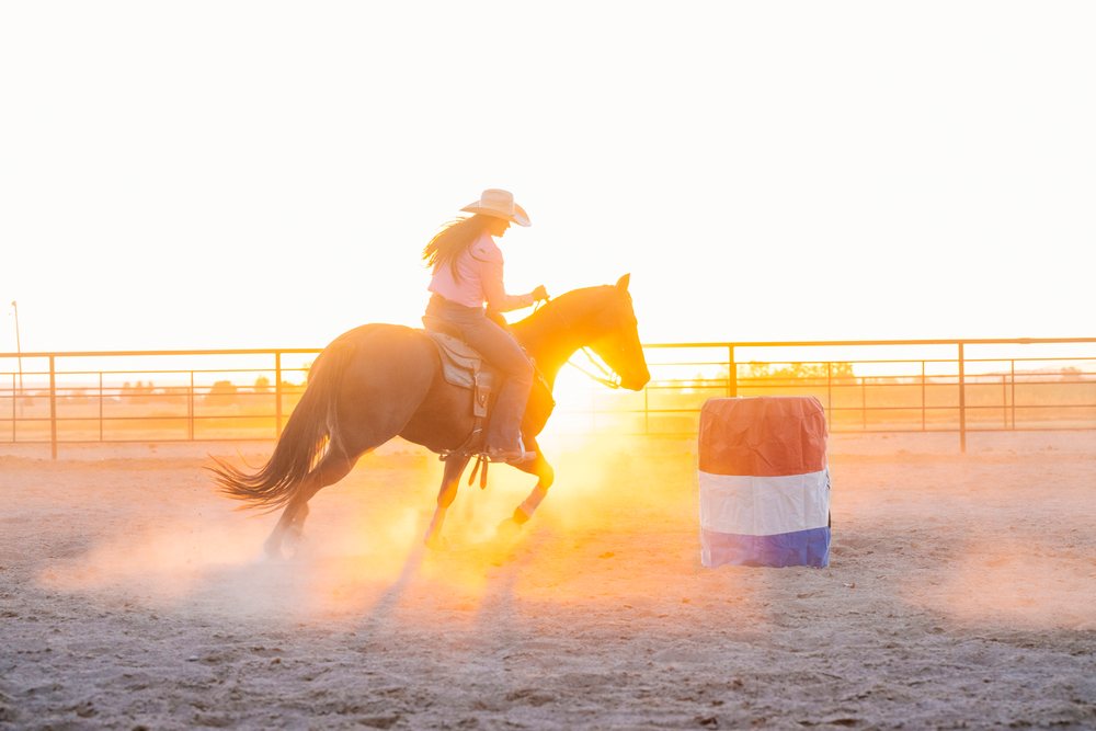 Horseback rider galloping around barrel in outdoor arena at sunset