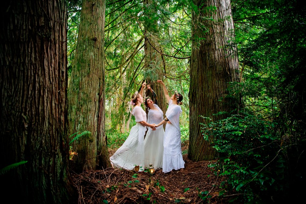Three women in white dresses holding hands in forest