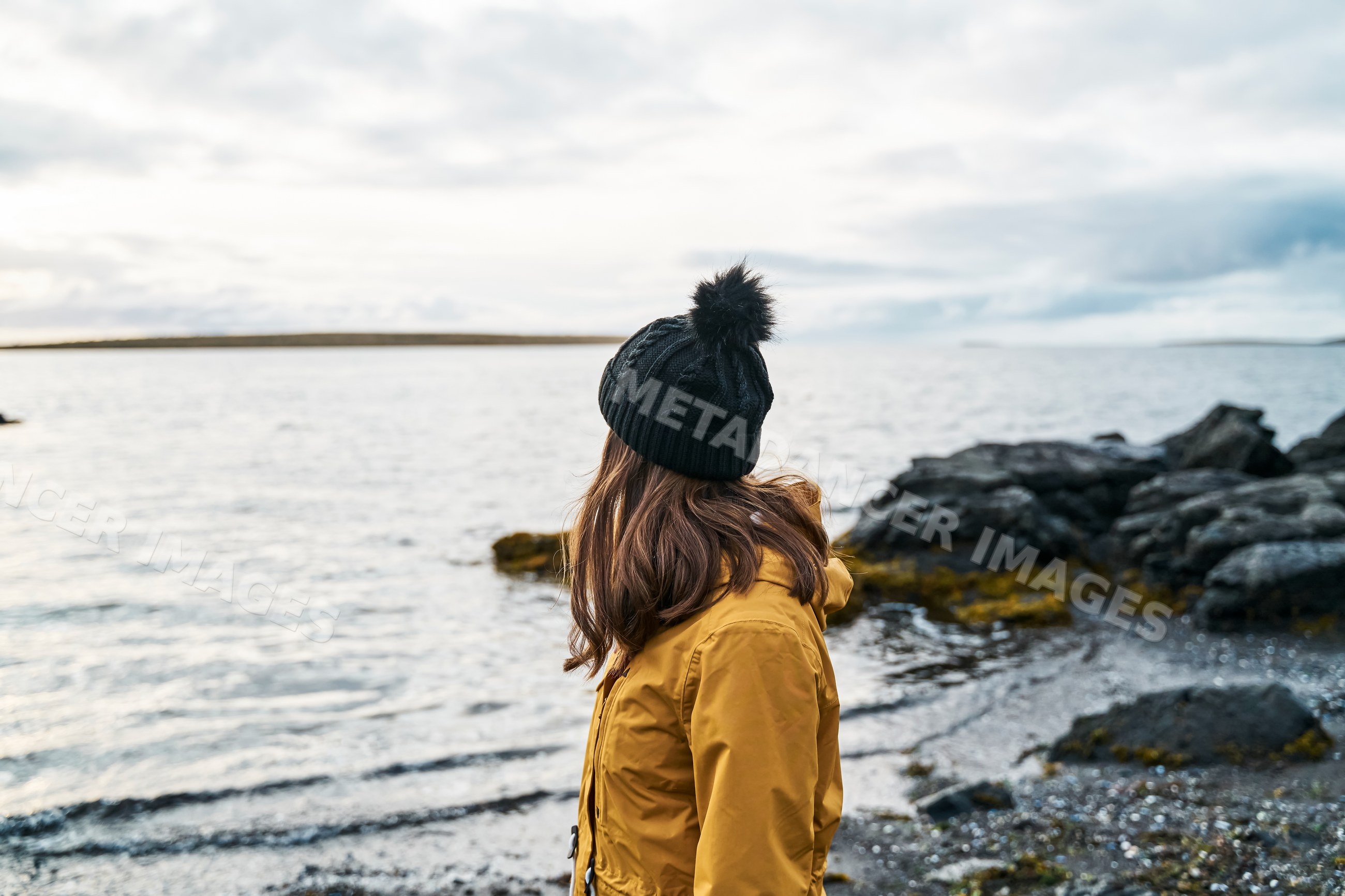 White Woman stands on rocky shoreline looking out over calm water