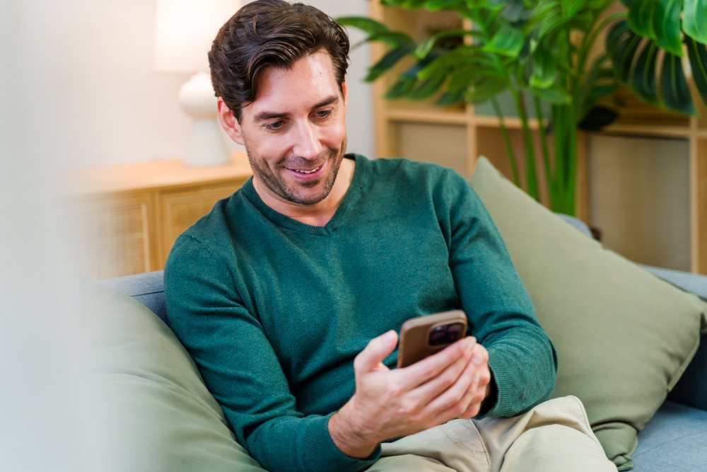White man holding smartphone sitting on couch in living room