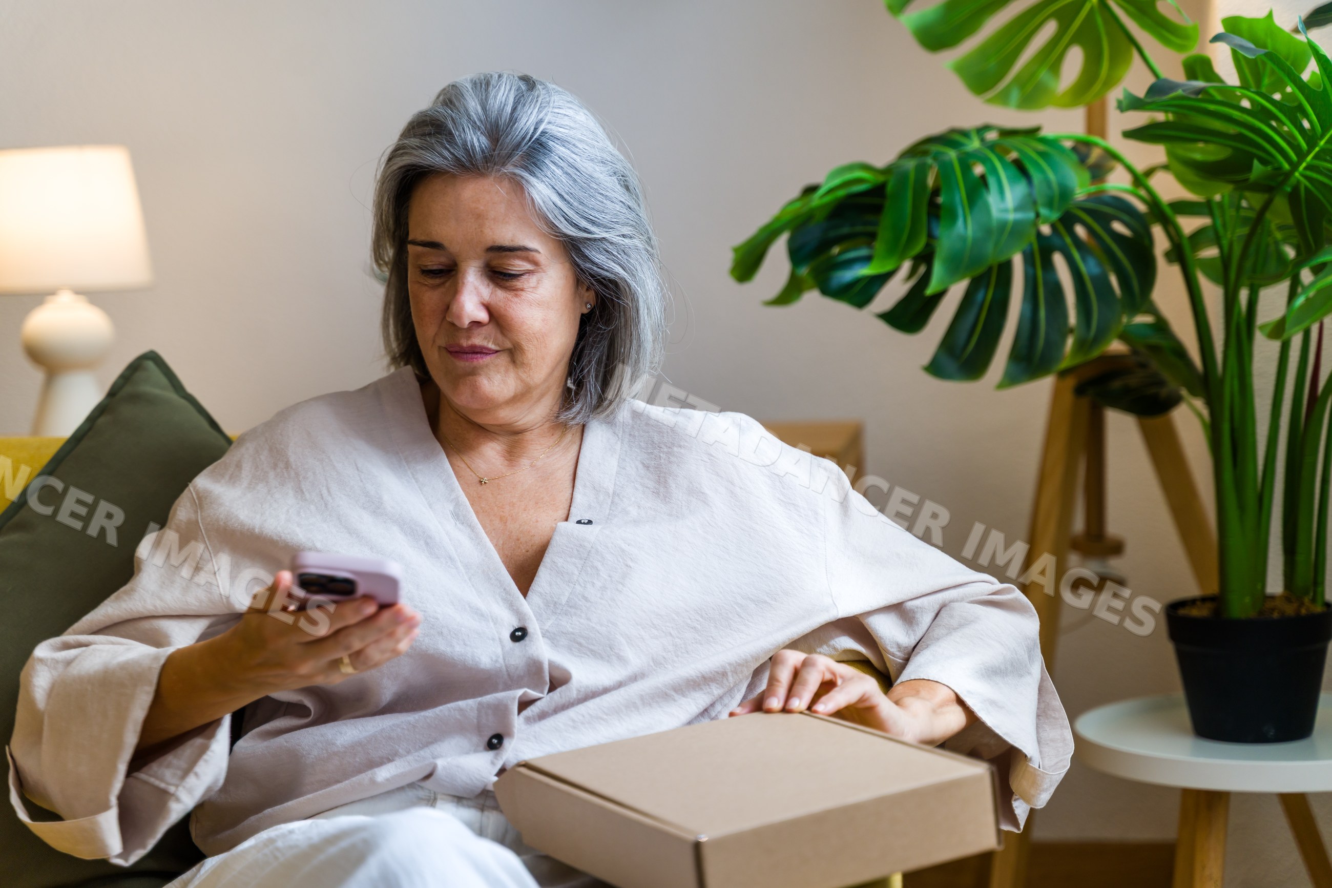 White woman sitting on sofa holding cardboard box using smartphone