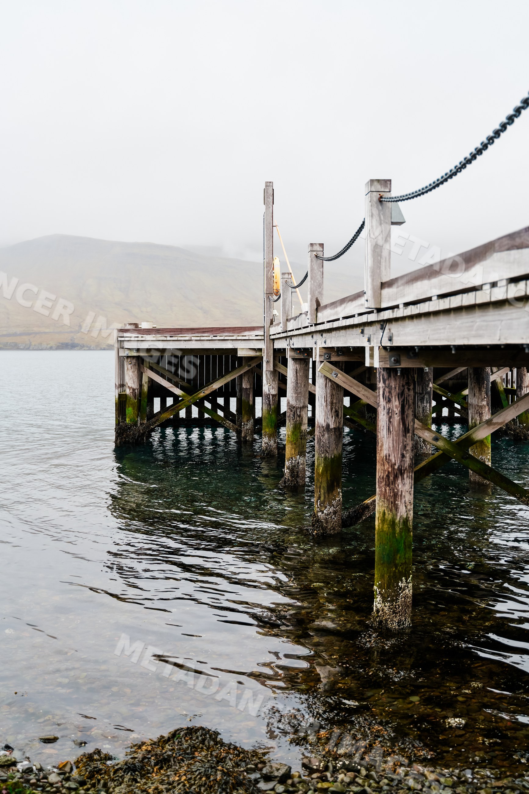 A wooden pier extends over calm sea water with a misty mountain