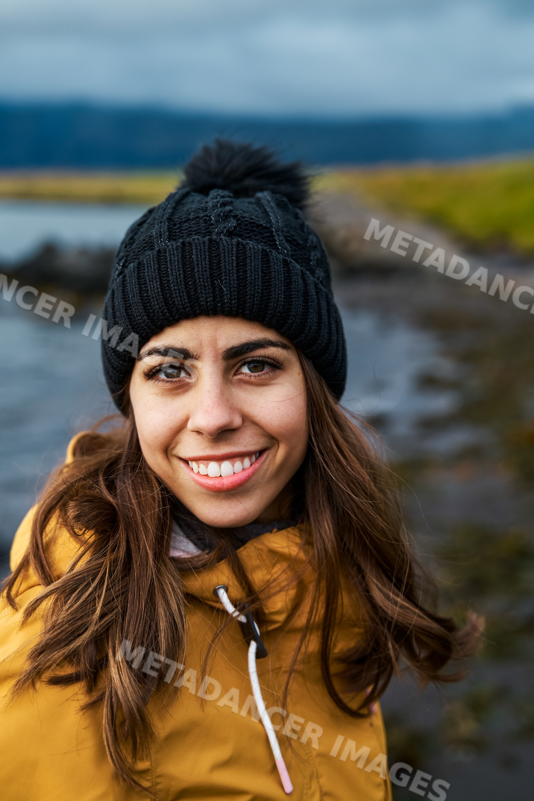A white woman smiles outdoors in cold weather near a rocky shore