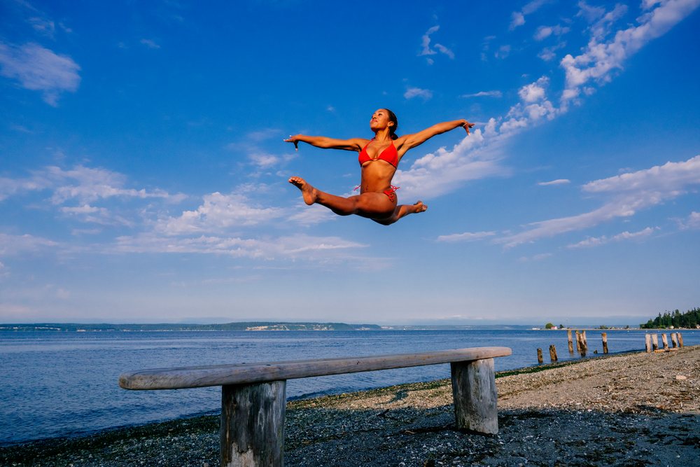 A woman jumps in a red swimsuit on a pebble beach under blue sky