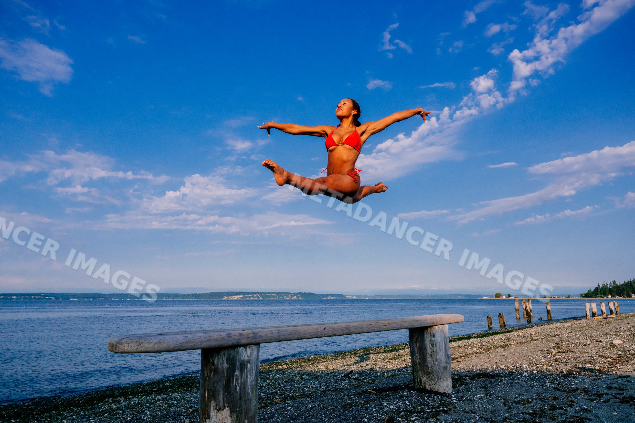 A woman jumps in a red swimsuit on a pebble beach under blue sky