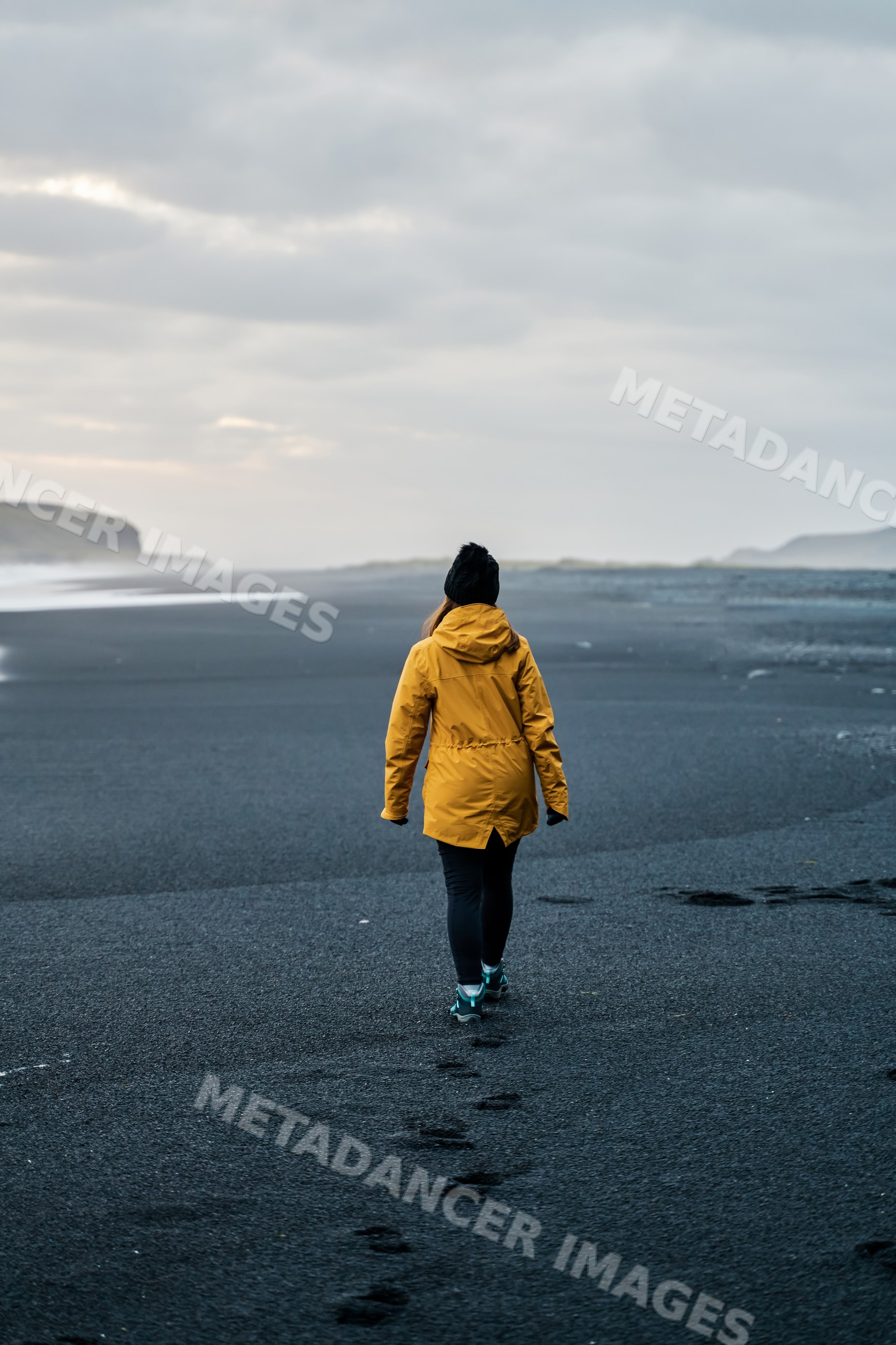 A white person walks on a black sand beach under an overcast sky
