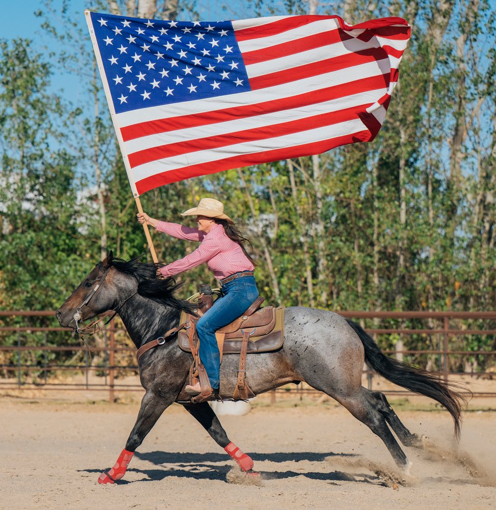 White woman riding horse holding american flag in outdoor arena