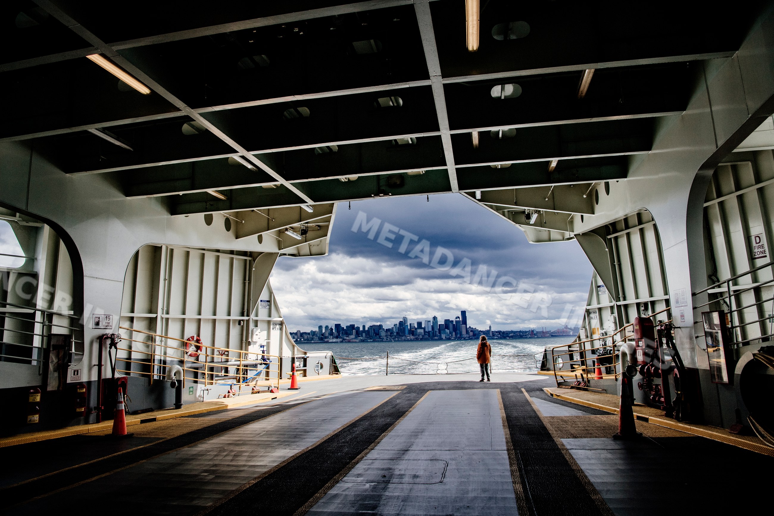 A person stands on a ferry deck viewing an urban skyline under cloudy