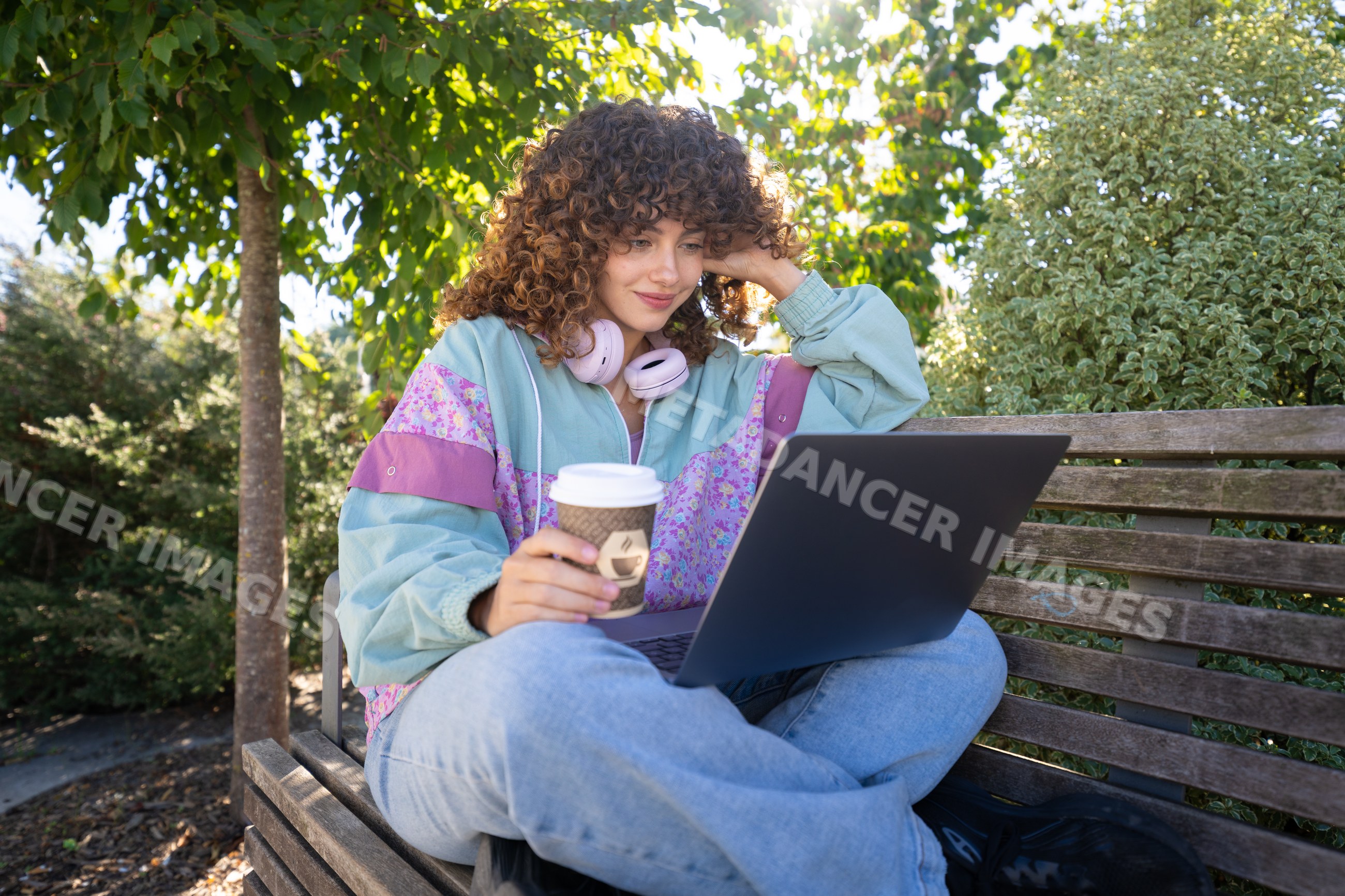 White woman using laptop on bench outdoors with coffee cup