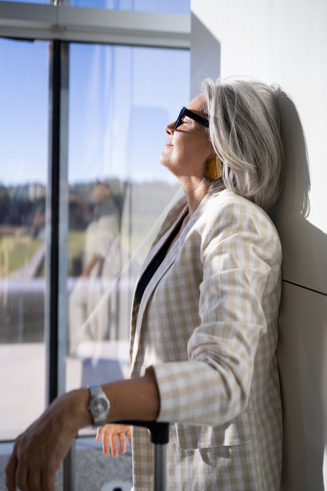 White woman with gray hair standing relaxed by window