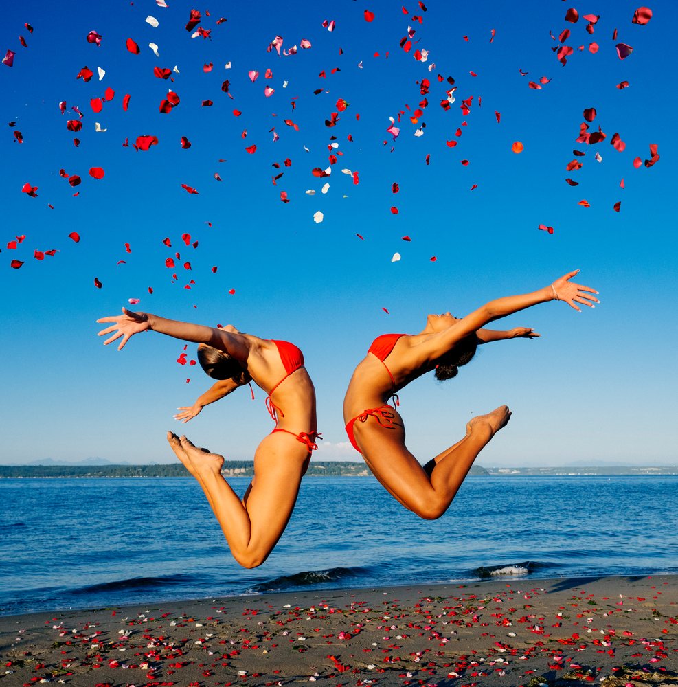Two women in red swimwear jump on a beach with rose petals in wa USA