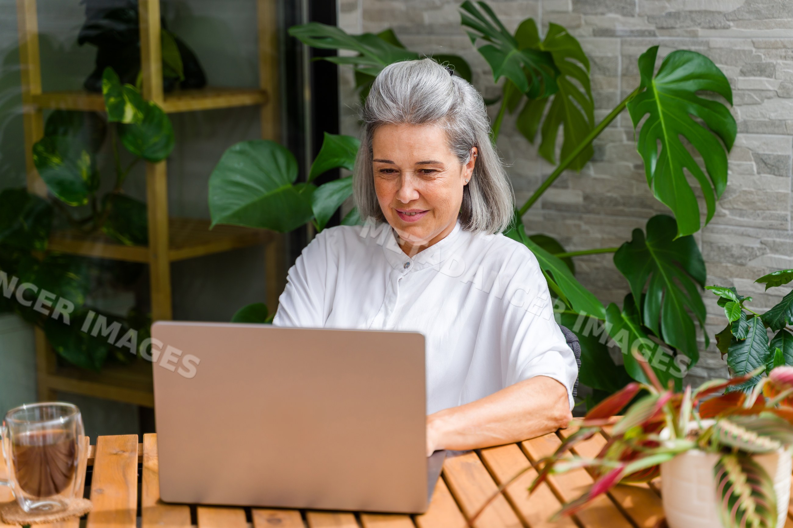 Mature white woman working on laptop in outdoor workspace with plants