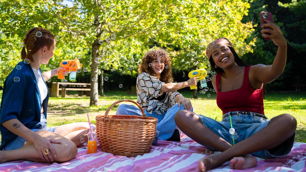 Friends playing with bubble guns and taking selfie on picnic blanket