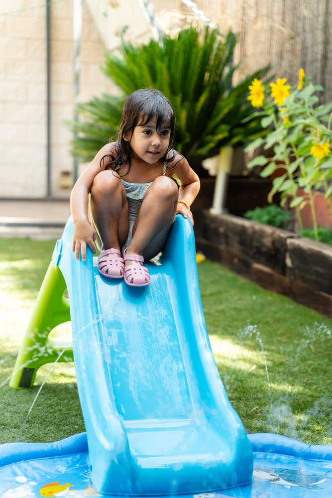A little white girl sitting on a blue water slide in a backyard garden