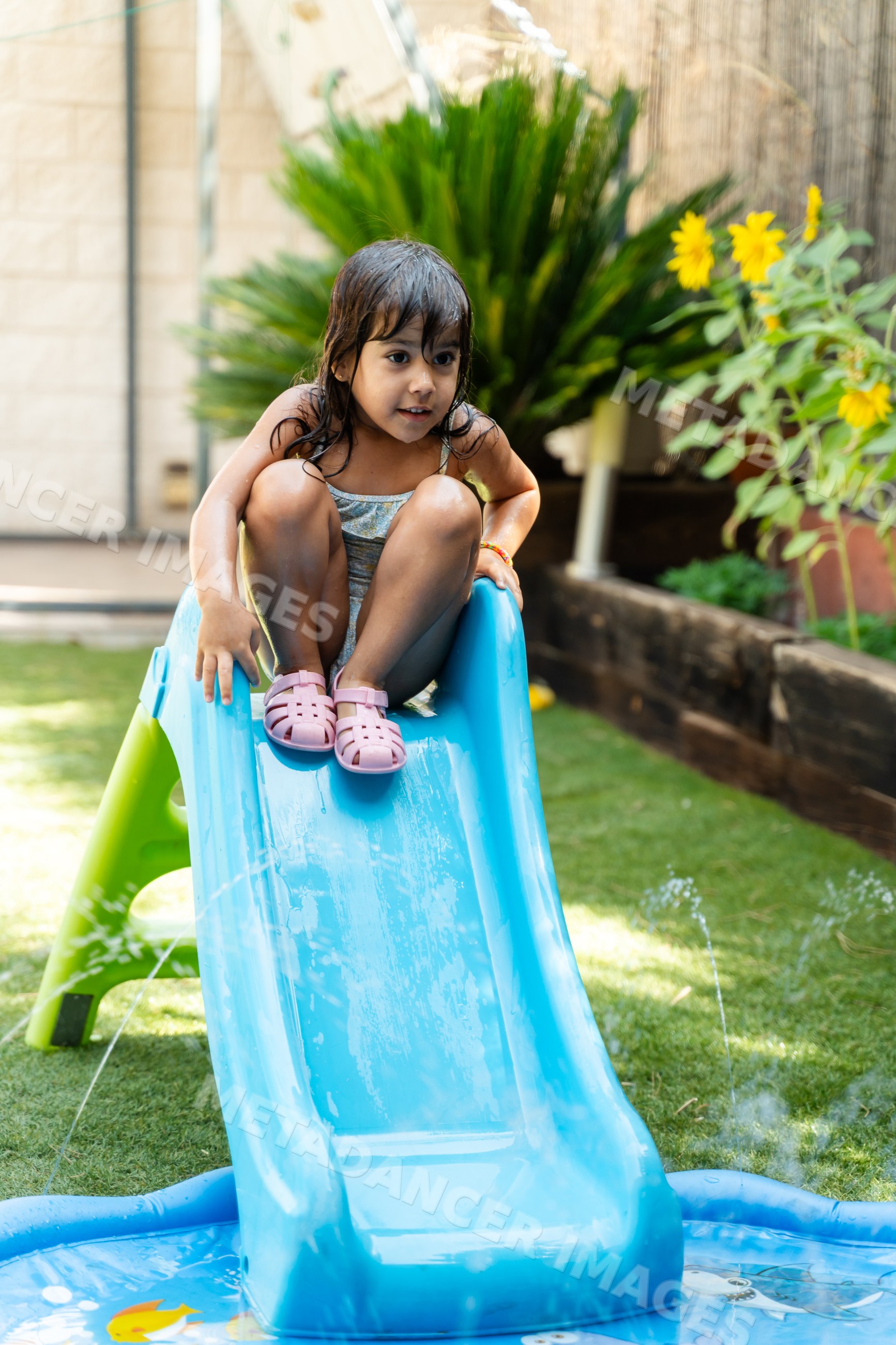A little white girl sitting on a blue water slide in a backyard garden