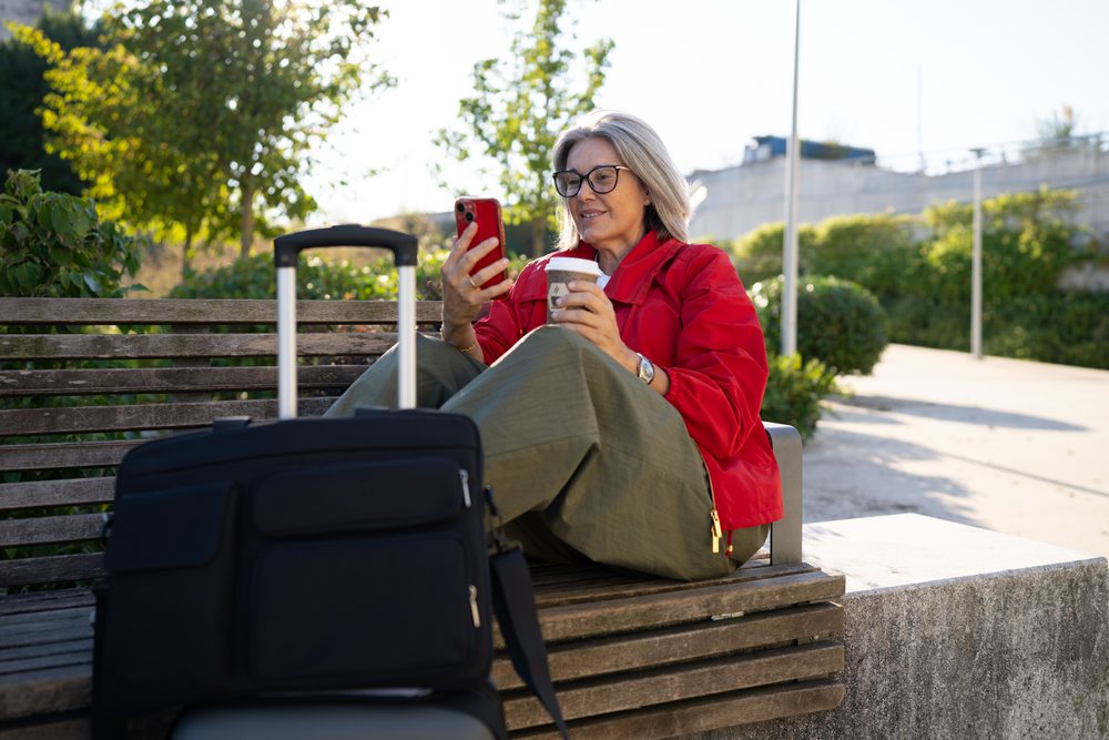White woman sitting on bench using smartphone with coffee and luggage