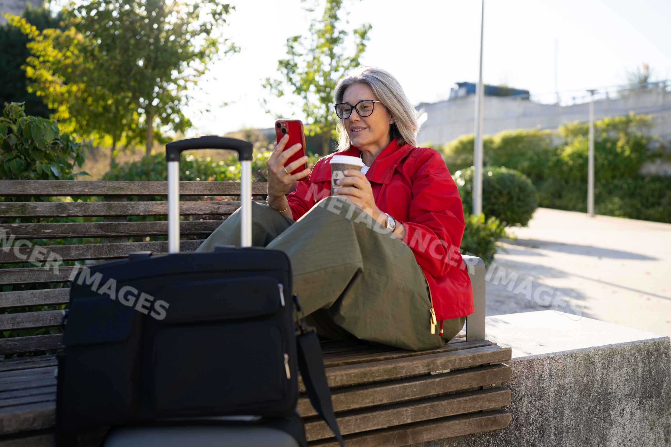 White woman sitting on bench using smartphone with coffee and luggage