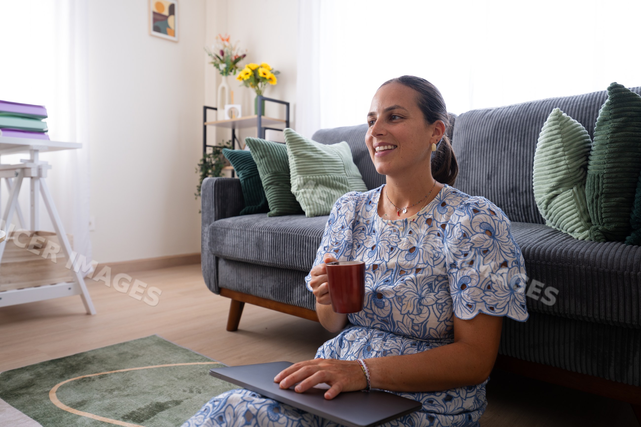 White woman sitting on floor with laptop and cup in bright living room