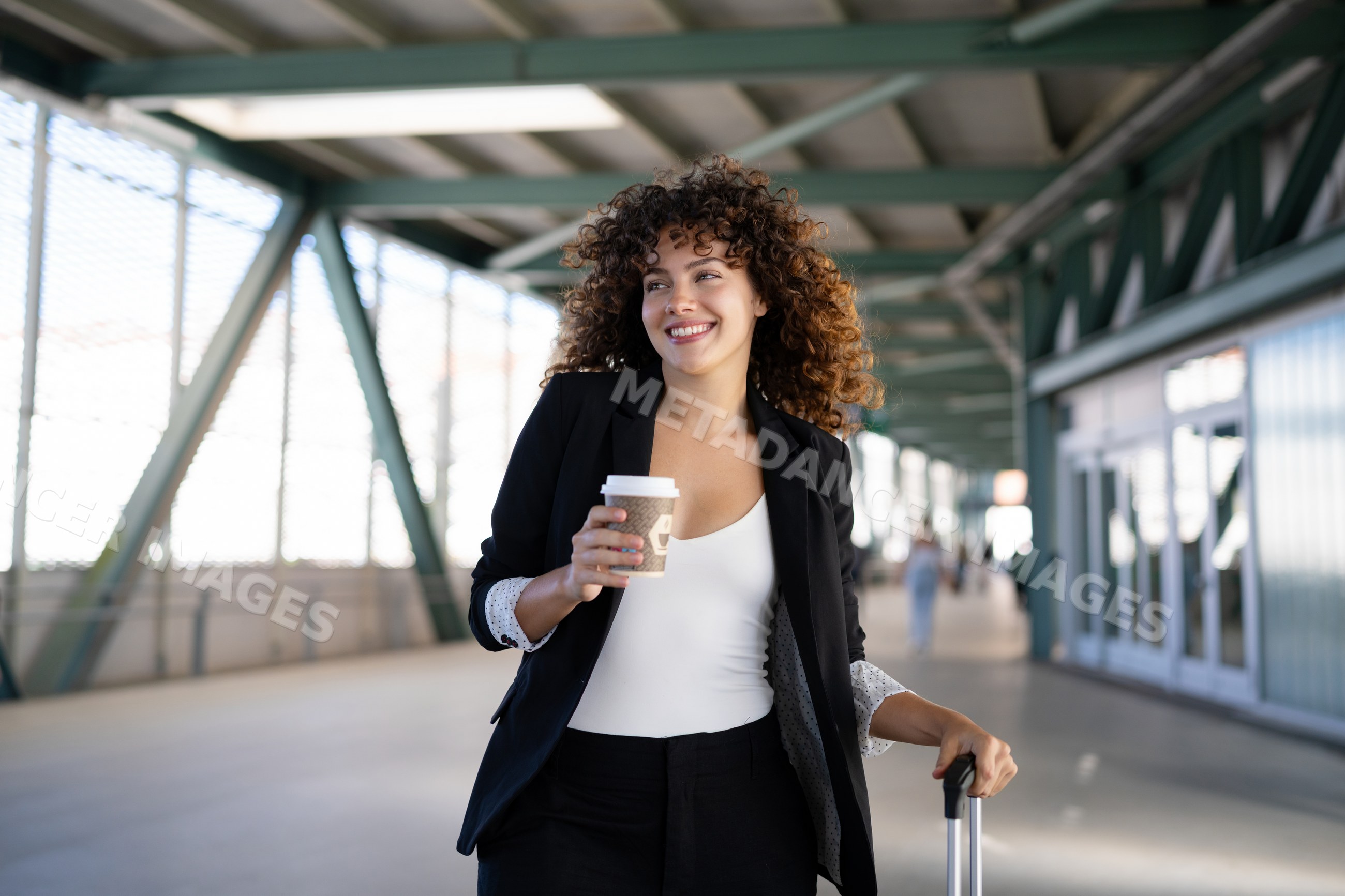Smiling white woman walking with coffee and luggage in corridor