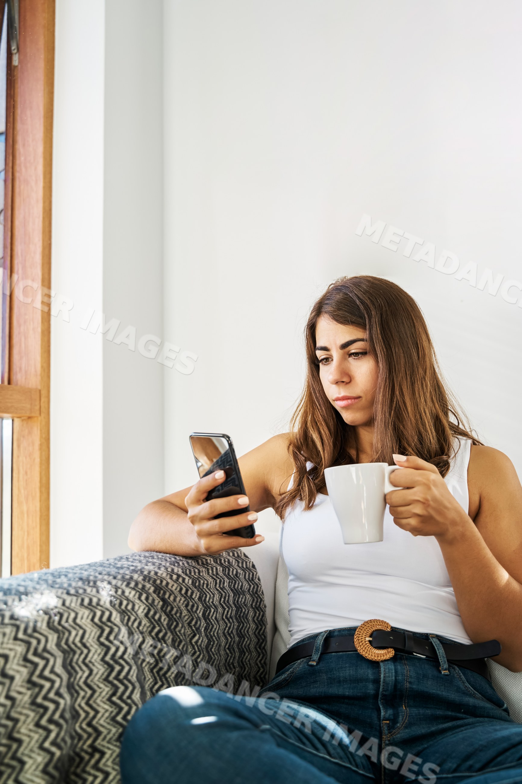 A white woman relaxes on a couch using a smartphone and holding
