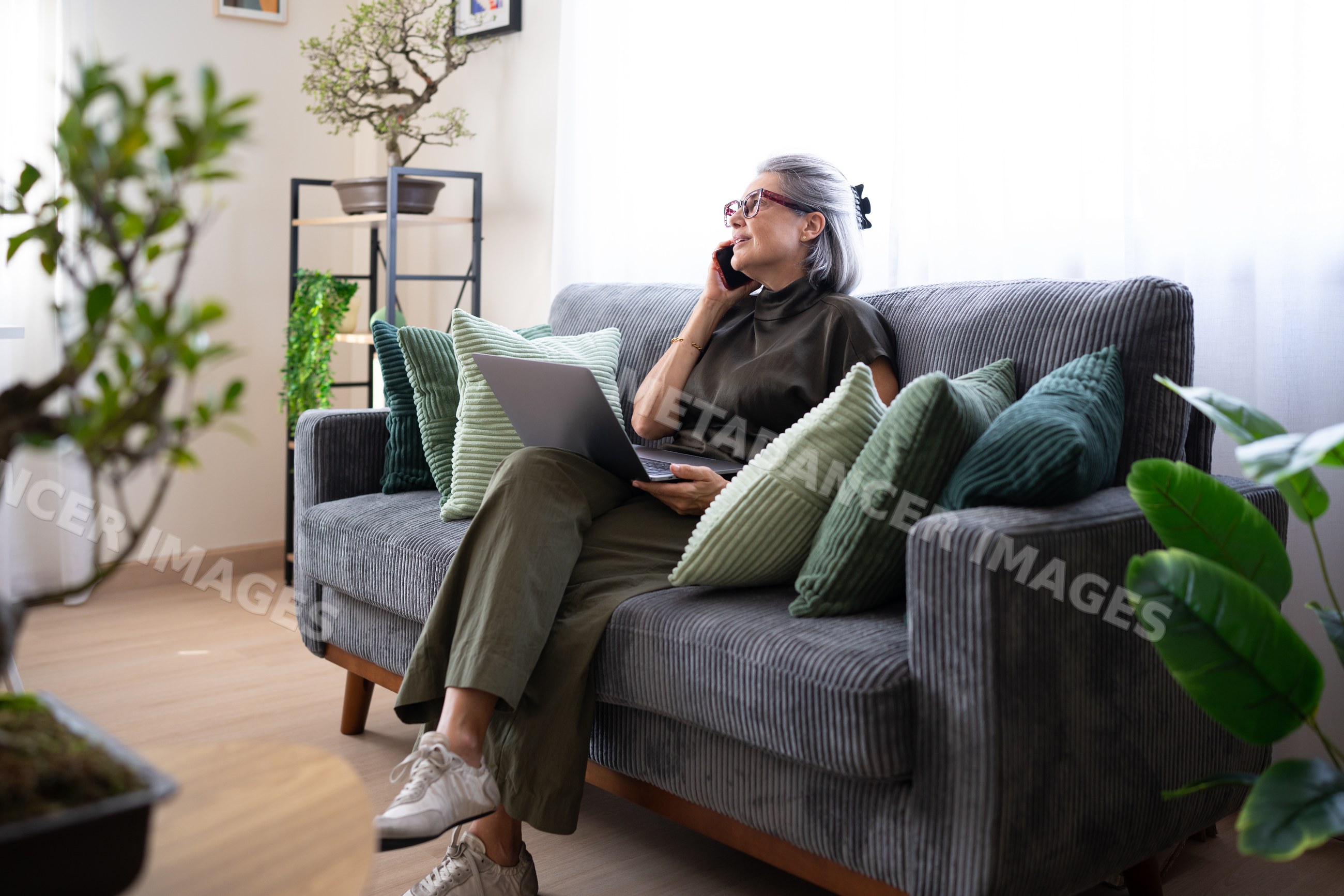 Senior white woman calling on phone and using laptop on gray sofa