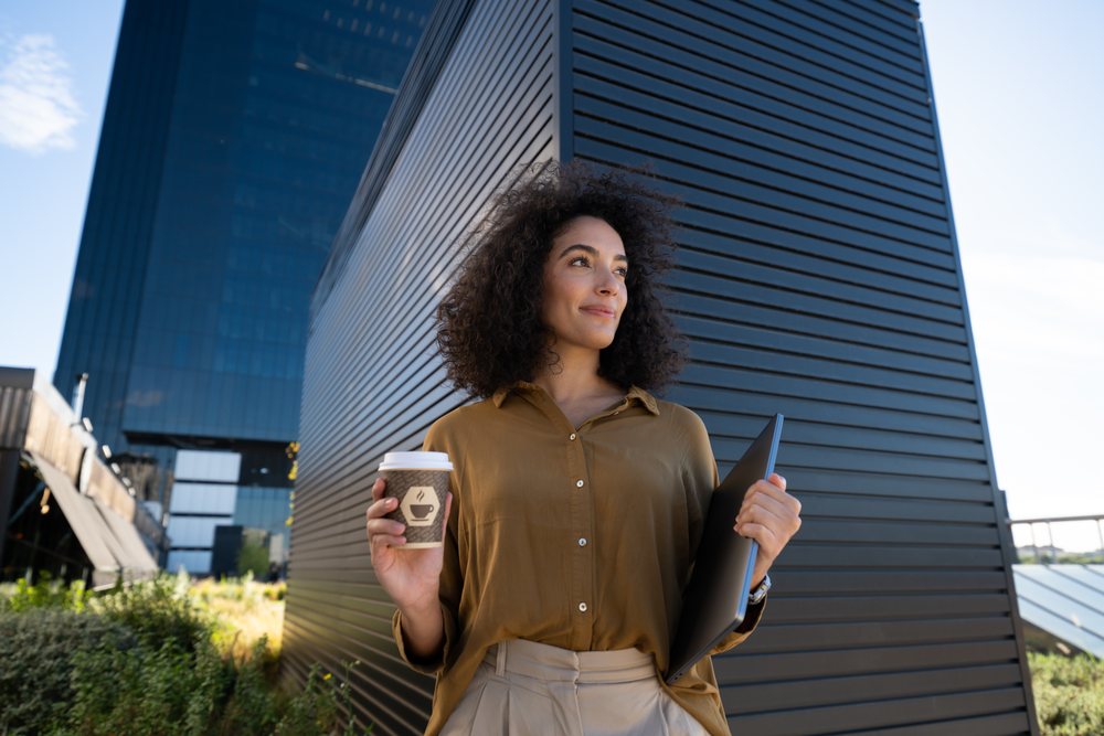 Woman holding coffee cup and laptop near modern building outdoors