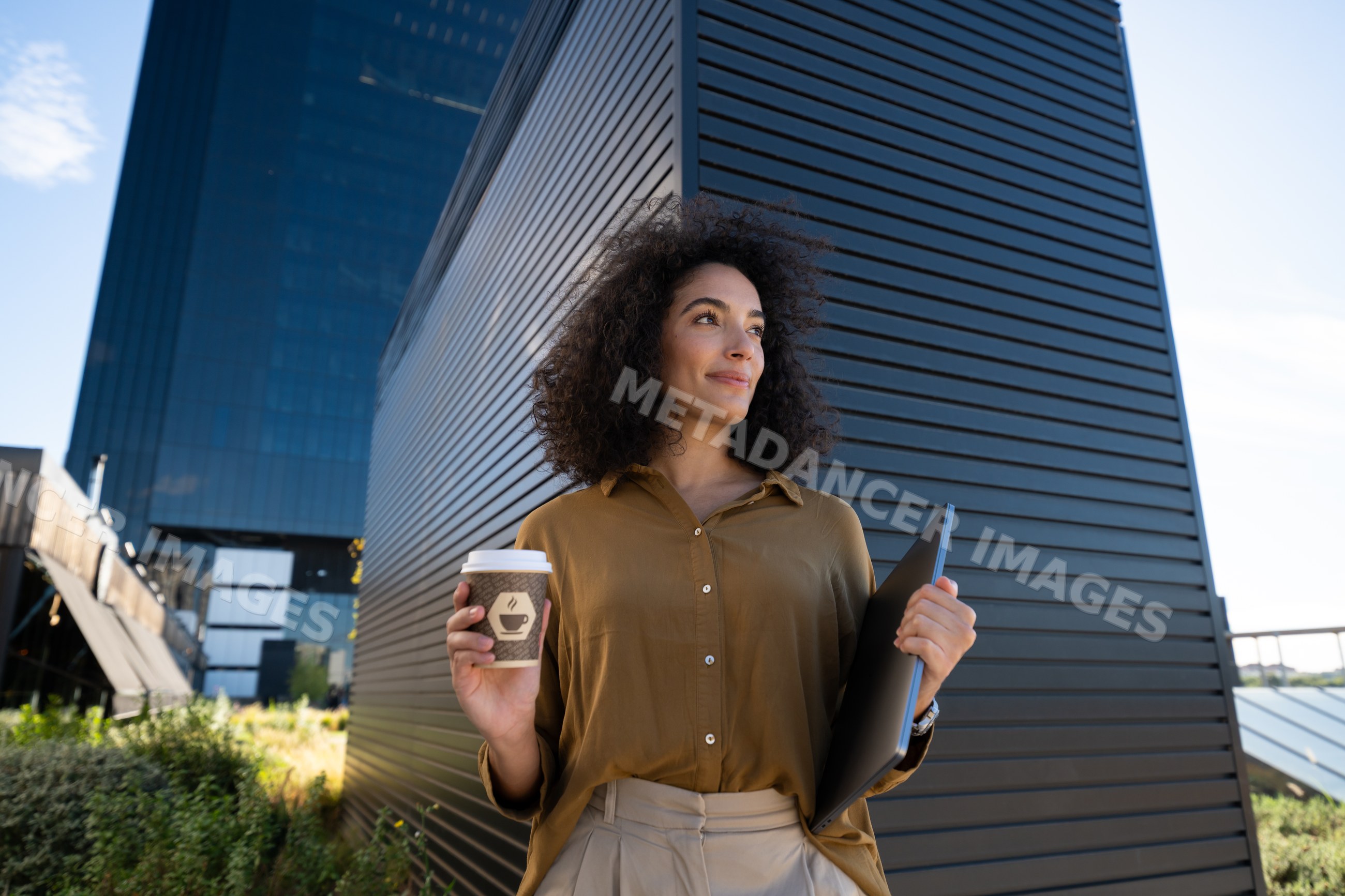 Woman holding coffee cup and laptop near modern building outdoors
