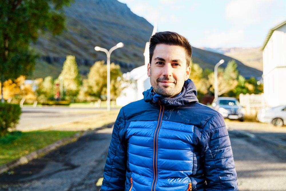 A young white man stands outdoors wearing a blue jacket in a street
