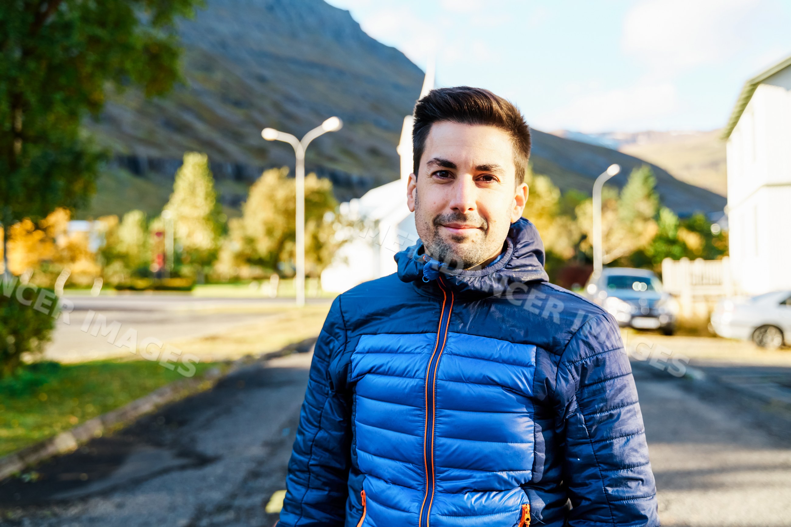 A young white man stands outdoors wearing a blue jacket in a street