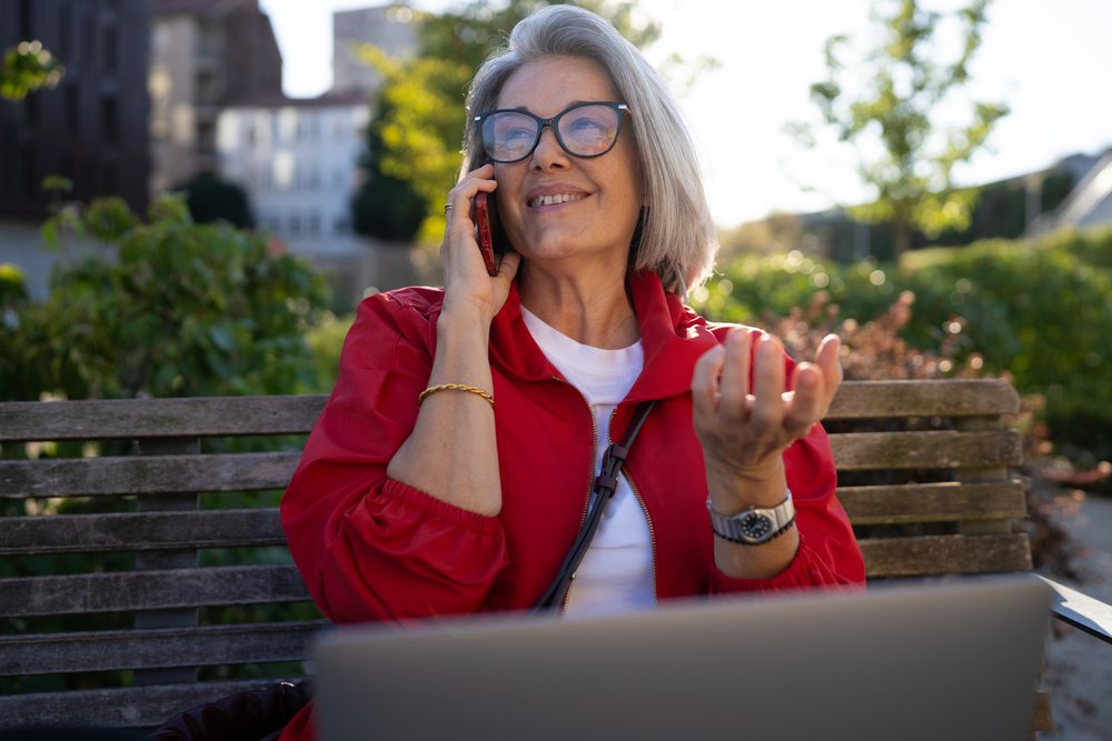 White woman talking on phone outdoors sitting on a bench with laptop